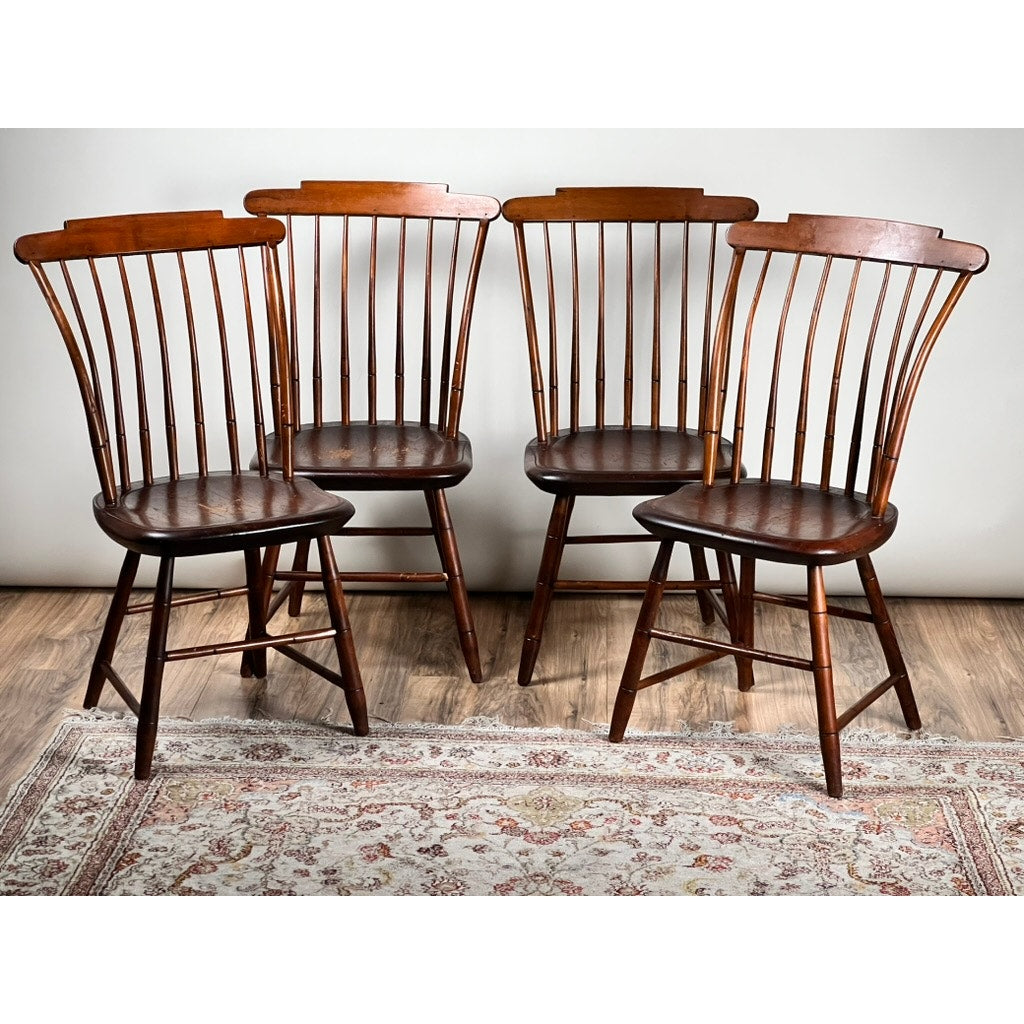 An Antique Set of Four New Hampshire Step Down Windsor Chairs by JR Hunt (early 19th c.) is arranged in a semi-circle on a light-patterned rug atop a wooden floor, against a plain light gray wall.