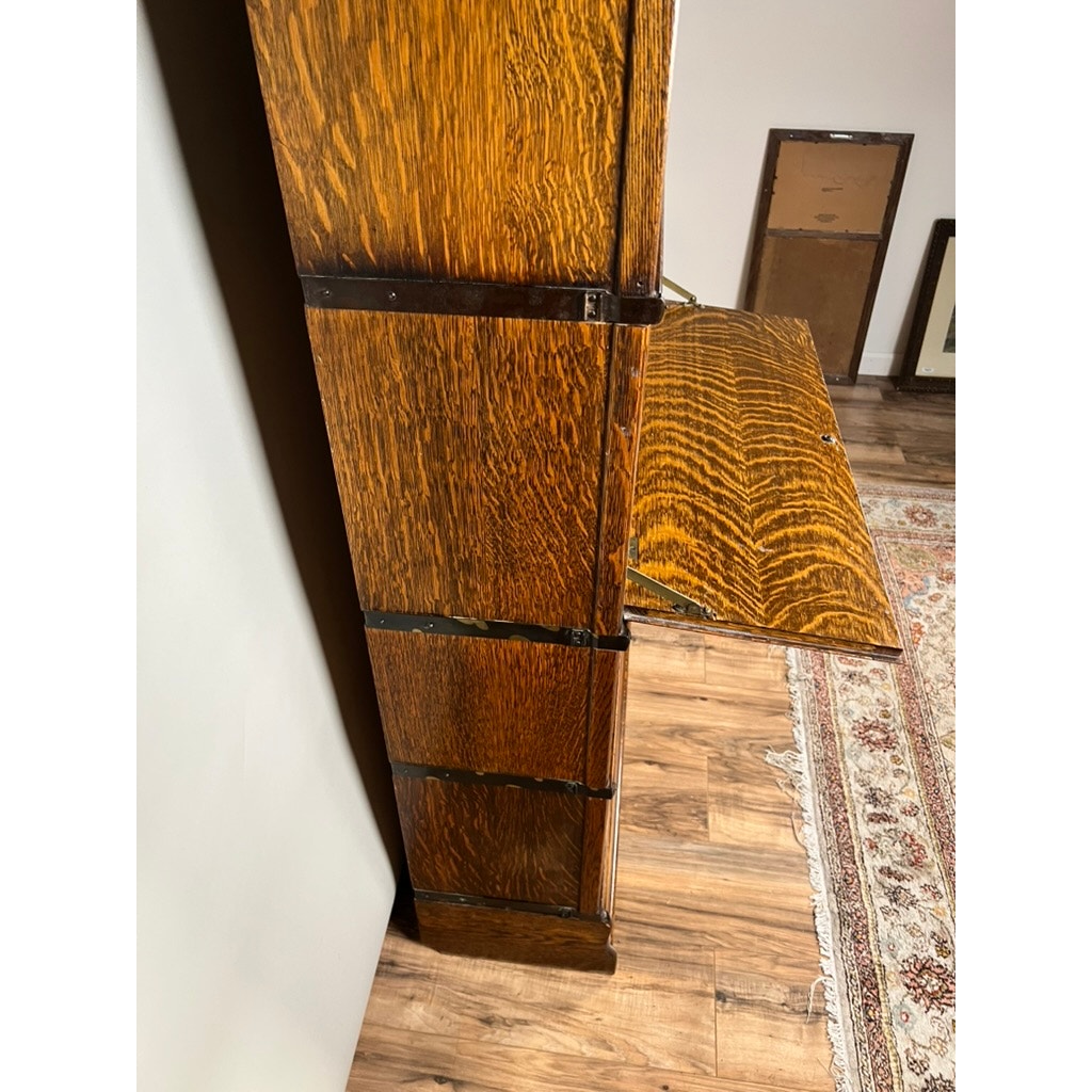 A partially open drop-down door reveals the secretary desk feature of this Antique Globe-Wernicke Quarter Sawn Oak 6-Stack Barrister Bookcase Secretary Desk (c. 1905–1915) by Globe Wernicke, standing on a wooden floor beside a patterned rug. Several framed items rest against the wall in the background, enhancing the timeless charm of this quarter-sawn oak classic.