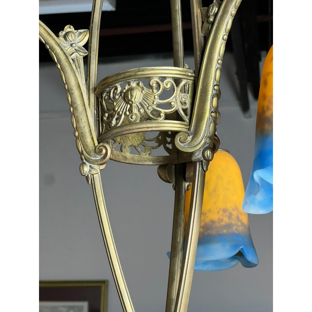 Close-up of a Vintage Muller Frères Lunéville French Brass and Art Glass Four Light Chandelier, showcasing ornate brasswork and Art Nouveau shades with blue-to-amber glass. A blurred picture frame is visible in the background.