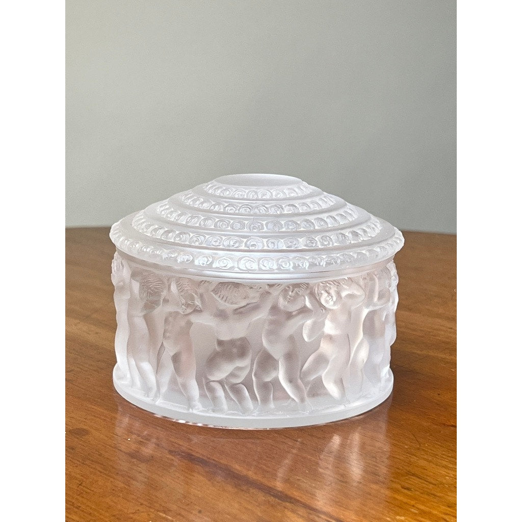 The Vintage Lalique Crystal Enfants Box by Lalique features frosted glass with reliefs of dancing nude figures encircling its sides, shown on a wooden surface against a plain gray background—a striking example of French art glass.