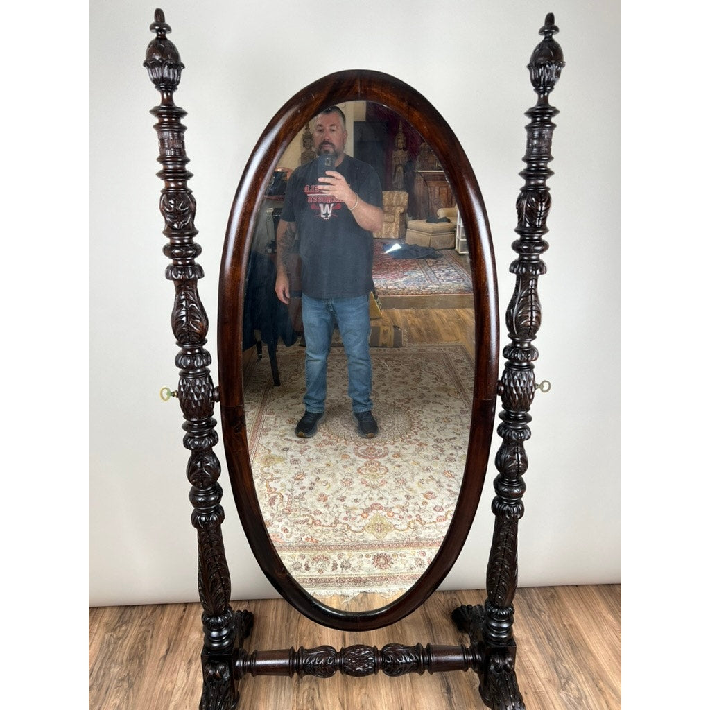 A man taking a selfie is reflected in an Impressive Antique American Empire Acanthus and Barbola Rose Carved Cheval Mirror c. 1840, set on a wooden floor with a patterned rug visible in the background.