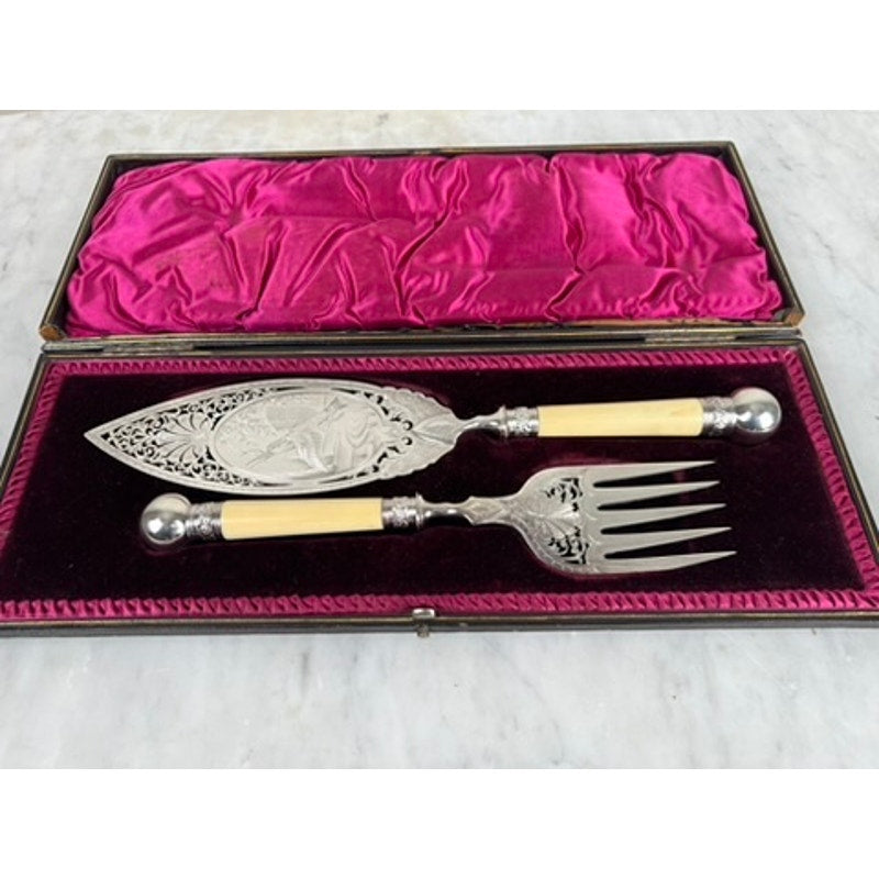 The Antique Victorian Silver-Plated Fish Serving Set includes a serving knife and fork with ivory-colored handles, beautifully presented in its original maroon velvet and satin box. Brand: None/Unknown.