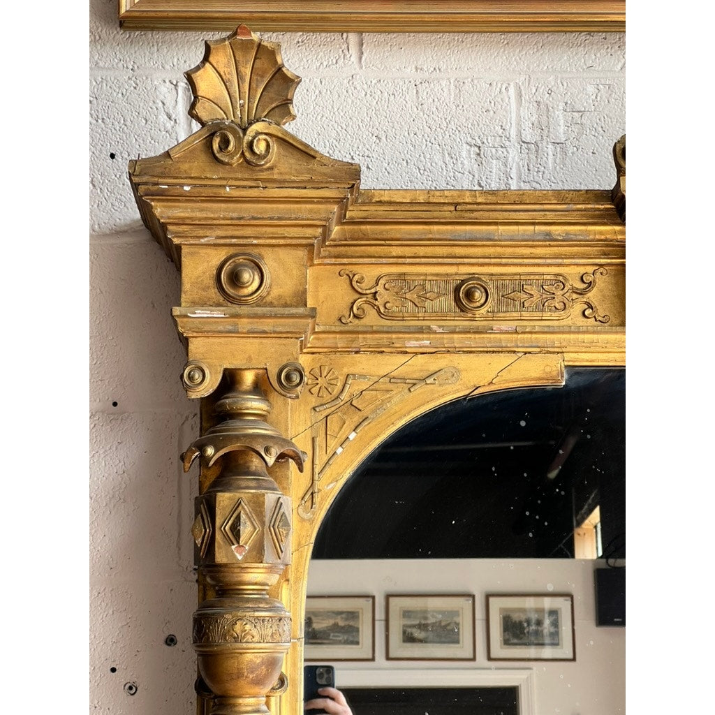 Close-up of an ornate, gold antique mirror frame with intricate Renaissance Revival carvings. This Exceptional Monumental Victorian Renaissance Revival Pier Mirror, circa 1870–1885, features a richly detailed giltwood frame. The reflection captures framed artwork on a white wall and part of a hand holding a phone to take the photo. Brand unknown.