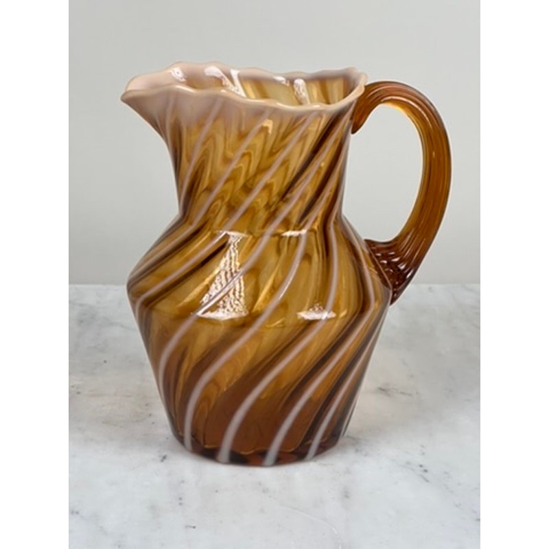 A Vintage Fenton Autumn Gold Opalescent Glass Spiral Pattern Pitcher with a scalloped rim and curved handle rests on a light marble surface. Brand is unknown.