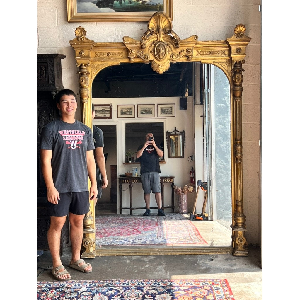 A smiling person stands before an Exceptional Monumental Victorian Renaissance Revival Pier Mirror, Circa 1870–1885; the reflection of another individual holding a phone appears in the ornate giltwood frame. The scene unfolds in a room adorned with artwork and patterned rugs, capturing the timeless elegance of Victorian Renaissance Revival style.