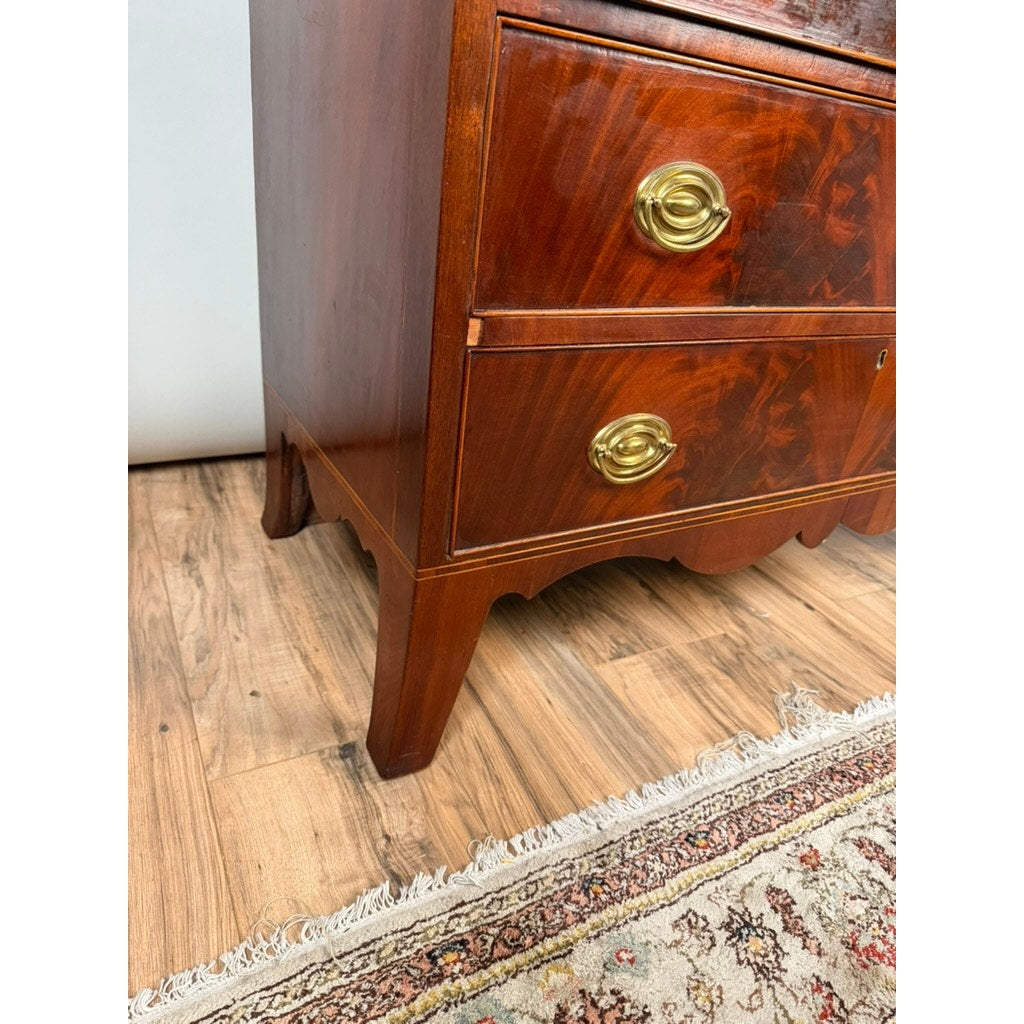 Close-up of the lower corner of a Federal Flame Mahogany Secretary Bookcase with Eagle Finial, c. 1810–1825, highlighting its rich flame mahogany grain and brass handles as it sits on a wooden floor next to a patterned rug with fringe.