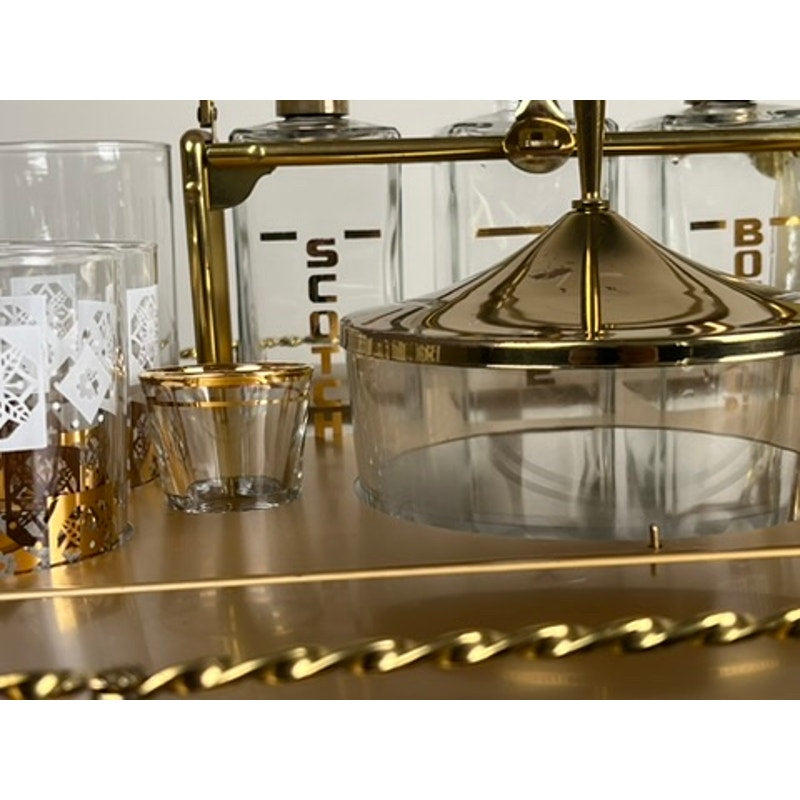 A close-up of the Vintage Mid Century Modern Bar Caddy c. 1960s displays gold-labeled Scotch and Bourbon decanters, patterned glasses, and a round glass container with a gold lid, arranged on a metallic vintage bar cart.