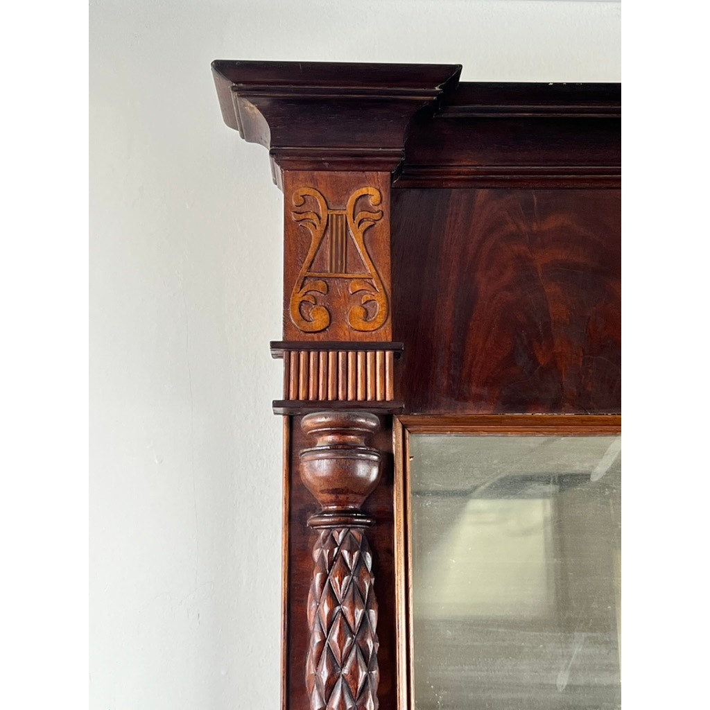 Close-up of the top corner of an Antique American Empire Mahogany Pier Mirror c. 1840, showing carved lyre motif, fluted accents, and twisted column design. Part of the glass mirror and white wall are visible. Brand unknown.