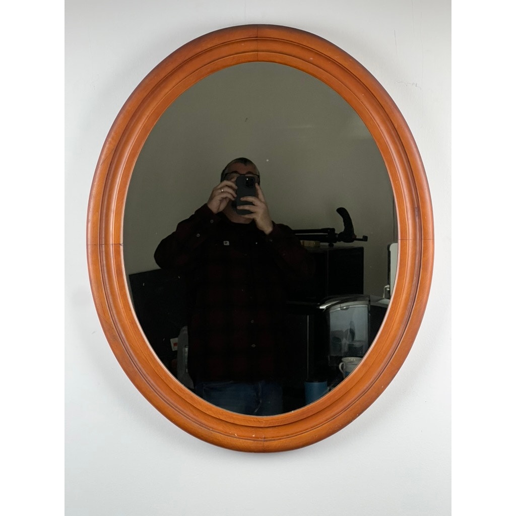 A person in a dark plaid shirt snaps their reflection in a Vintage Oval Maple Wall Mirror hanging on a white wall, with assorted background objects visible. Brand: None/Unknown.