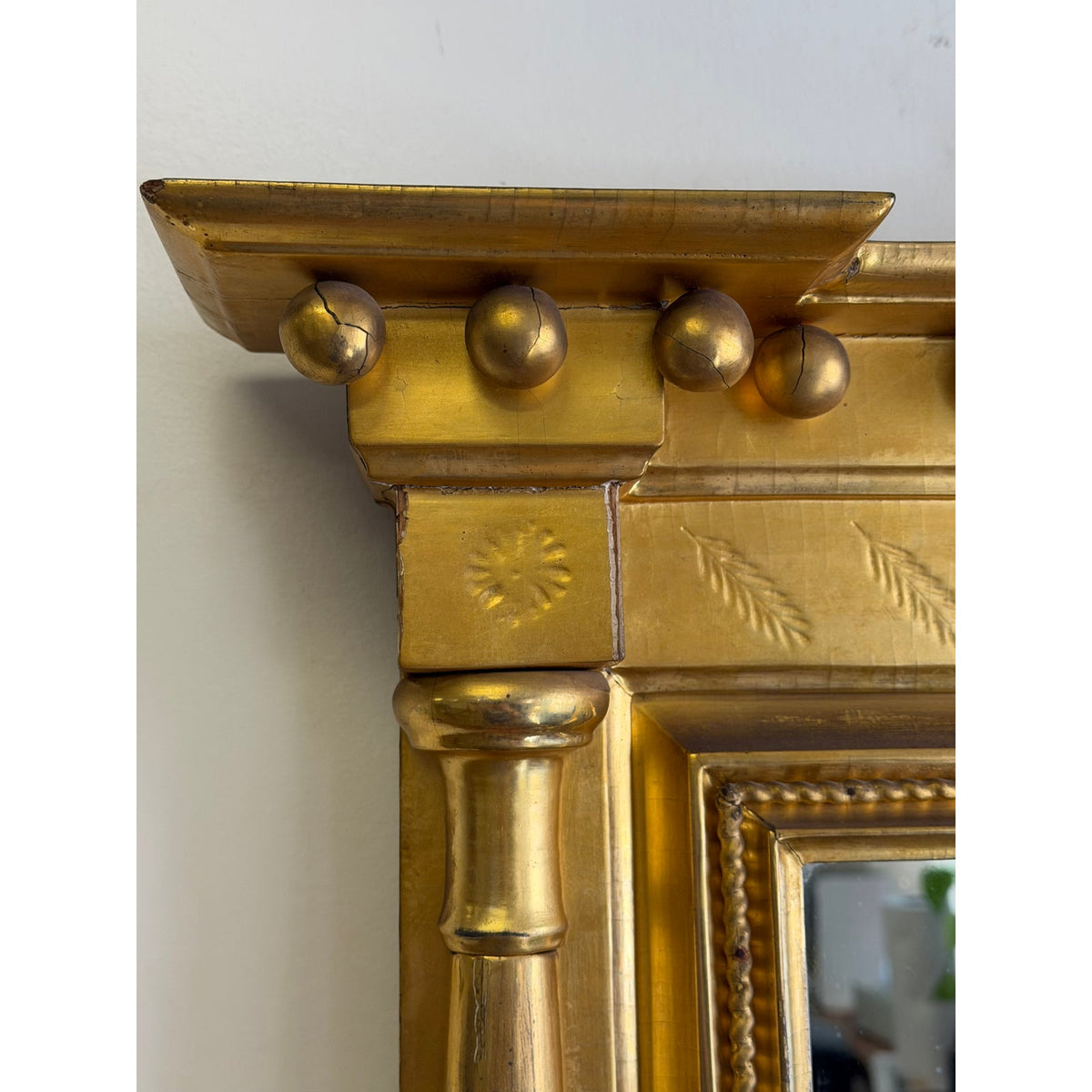 The upper corner of an American Federal Giltwood Two-Plate Looking Glass in gold-painted wood, featuring a cornice with spherical ornaments, a carved star motif on the column capital, and delicate leaf engravings along the top rail.