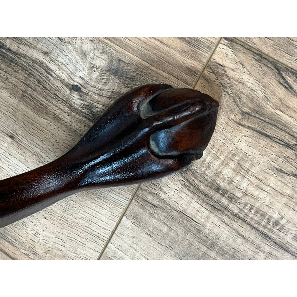 A close-up of a wooden cane handle intricately carved in the shape of an animal’s paw, resting on a wood-patterned floor beside a Pearl City Antiques Antique George III Tilt-top Tea Table from the early 19th century.