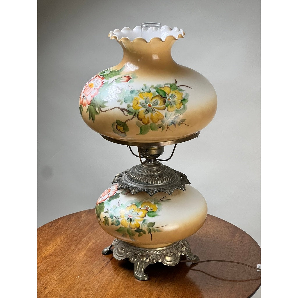 A Vintage Hand-Painted Floral Milk Glass Hurricane Parlor Lamp with ornate hardware and a scalloped top sits elegantly on a round wooden table. Brand: None and/or Unknown.
