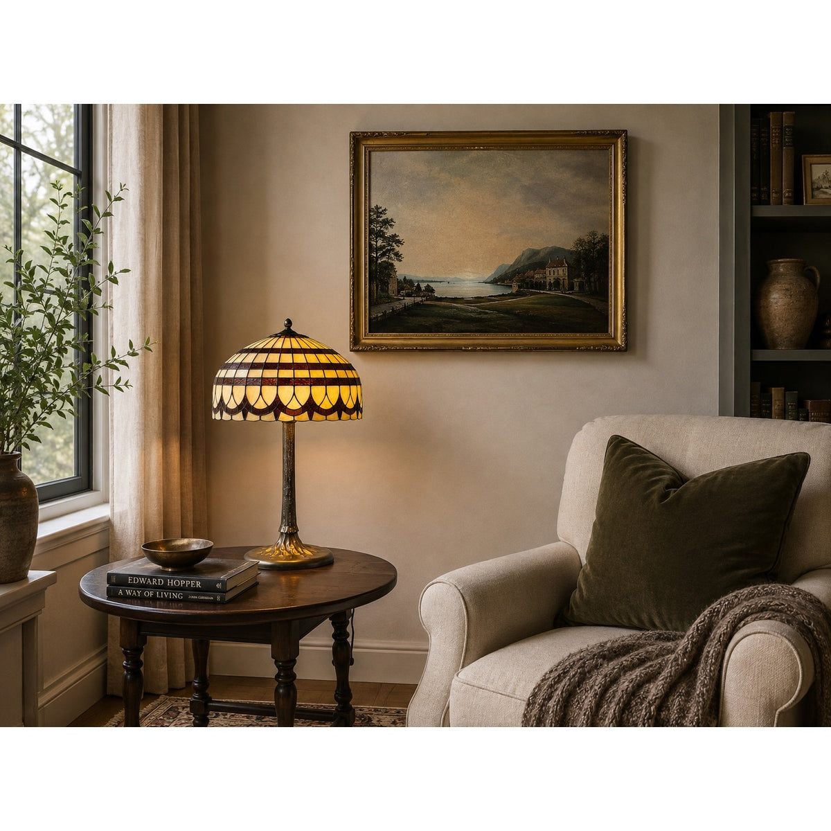 A cozy living room with an armchair, a dark round side table displaying the Pearl City Antiques Vintage Tiffany Revival Leaded Stained Glass Table Lamp with a cast brass tree-form base, books, a throw blanket, a plant by the window, and a framed landscape painting on the beige wall.