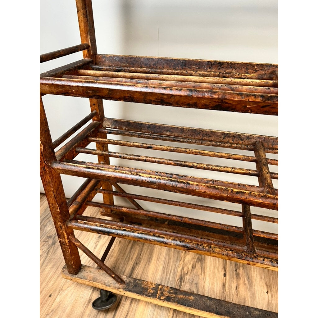 A close-up of an Antique Oak Shoe Factory Rolling Rack, c. late 19th century, with multiple slatted shelves and original industrial casters, showing signs of age and wear on a wood floor. Brand unknown.