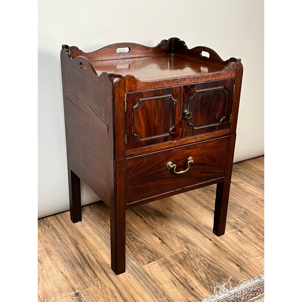 An Antique George III Bedside Commode with a pull-out leather surface from the late 18th century, featuring a scalloped edge, two cabinet doors, and a brass-handled drawer, shown on a wood floor beside a patterned rug.