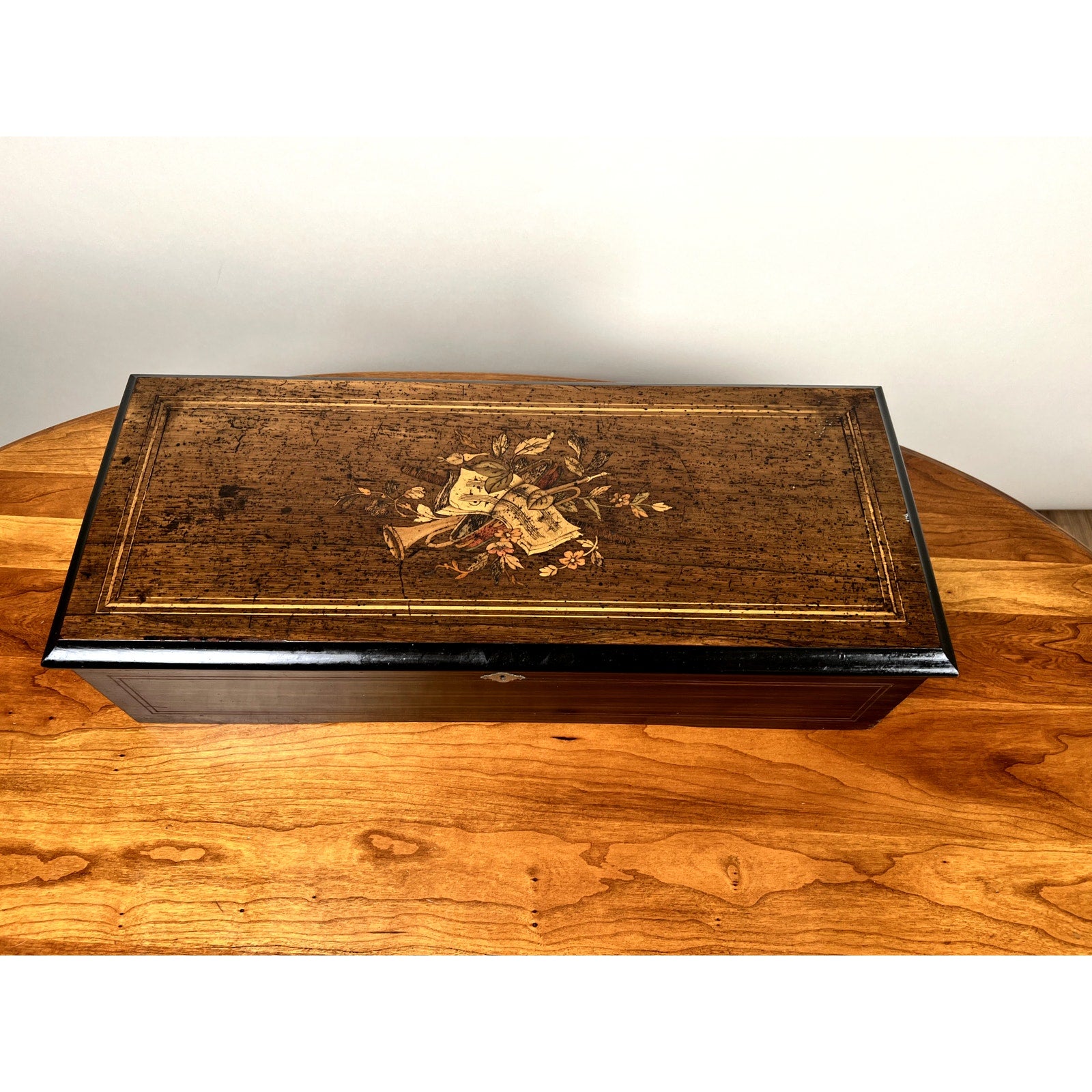 An Antique Swiss 12 Tune Music Box from the late 19th century, with a decorative floral and trumpet design on the lid, rests on a wooden table. Brand is unknown. The wooden box has a vintage, slightly worn finish.
