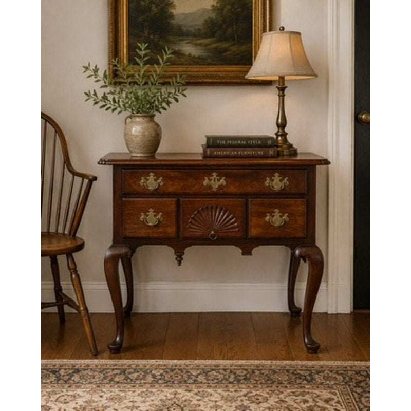 An 18th Century American Queen Anne Walnut Shell-Carved Lowboy (c. 1740–1770) with brass handles displays a lamp, two books, and a vase filled with greenery. A wooden chair sits beside it, while a framed landscape painting hangs above. The floor is adorned with a patterned rug.