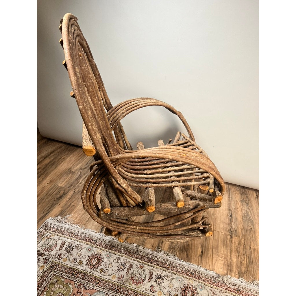 The Vintage Adirondack Bent Willow Arm Chair, crafted from curved branches in a rustic, artistic style, sits on a wooden floor beside a patterned rug and a plain white wall. Brand is unknown.