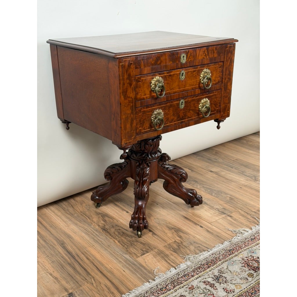 An American Classical Figured Mahogany Work Table, circa 1830–1845, featuring a rectangular top, two drawers with lion-head brass pulls, and a central pedestal base with carved feet.