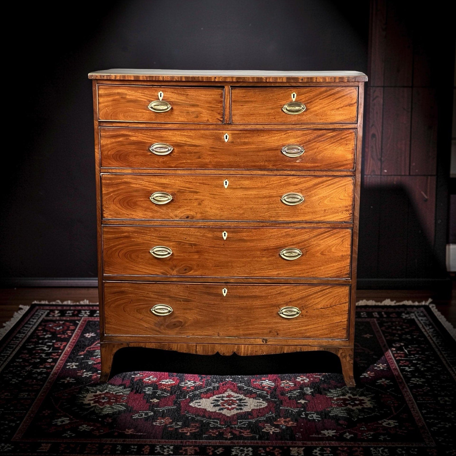 An Antique George III Two Over Four Chest with French Feet from the early 19th century stands on a patterned rug against a dark background, featuring six drawers, brass handles, and keyholes.