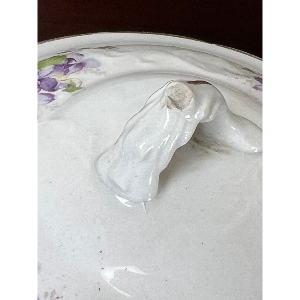 Close-up of a white ceramic lid with a curved handle and small hole, featuring purple floral patterns near the edge. This lid is part of the Antique Victorian Porcelain Wash Stand Set by Leland, signed and dating to the late 19th century.