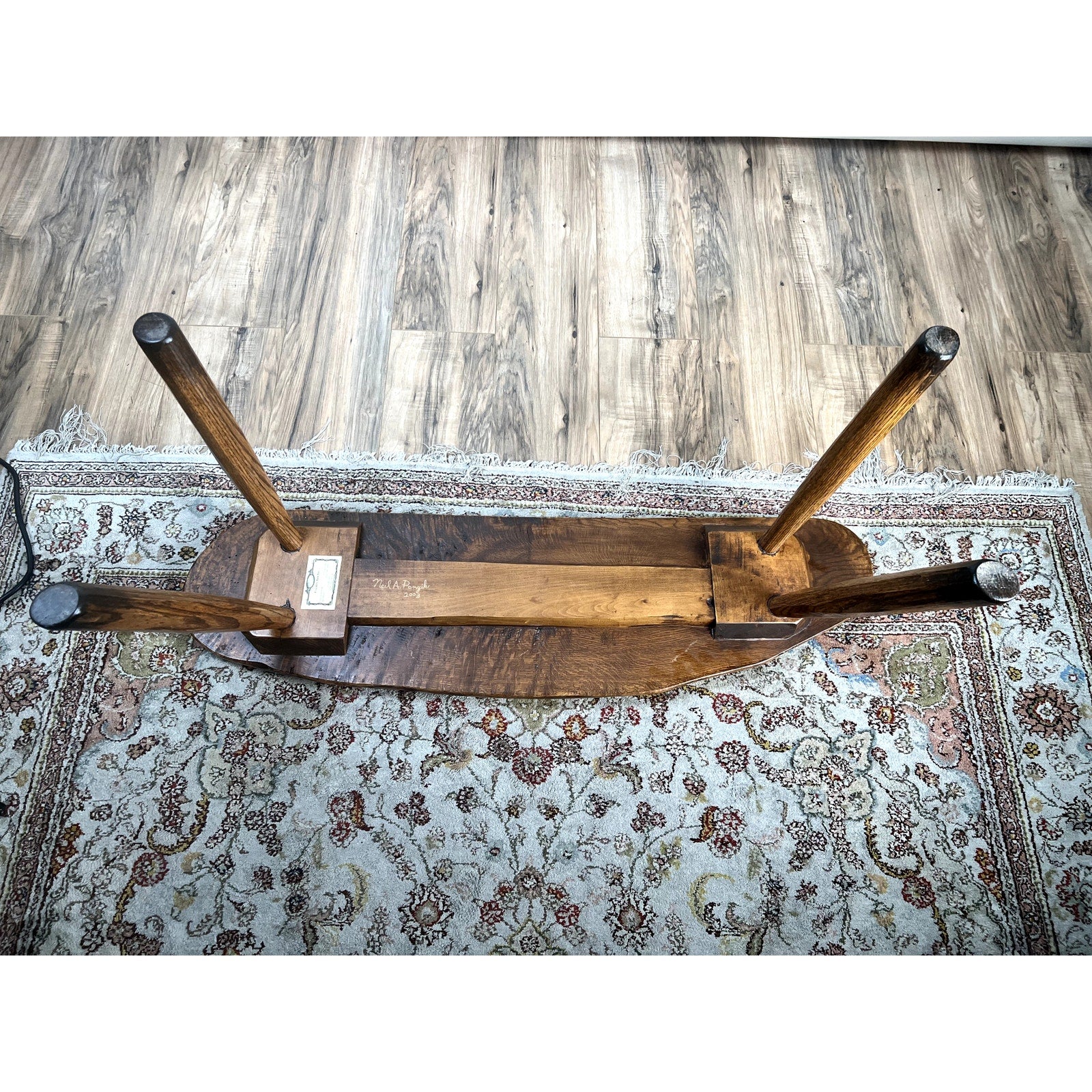 An overturned Vintage Hand Made Oak Wabi Sabi Bench signed Neil Panyik, showing its underside and four legs, rests on a patterned rug atop a wooden floor.