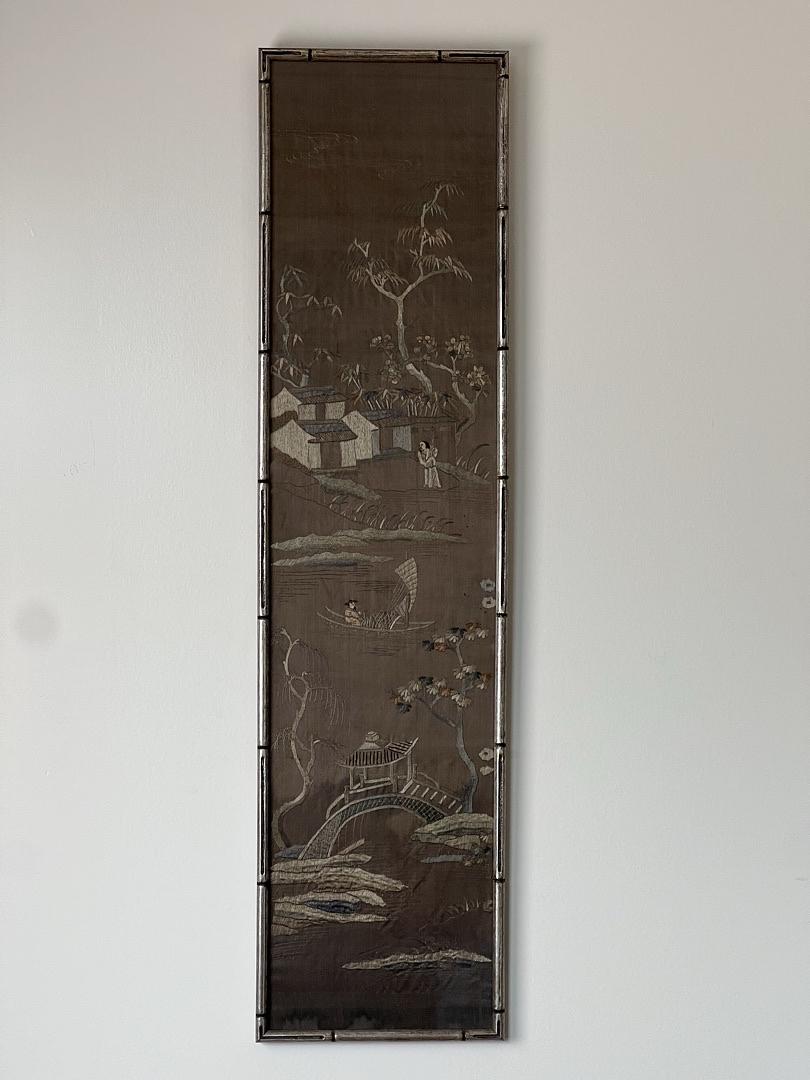 The Pearl City Antiques Antique Chinese Embroidered Panel on Silk (late 19th century) features a landscape with houses, trees, a boat, and mountains in a thin dark frame, displayed against a light wall.