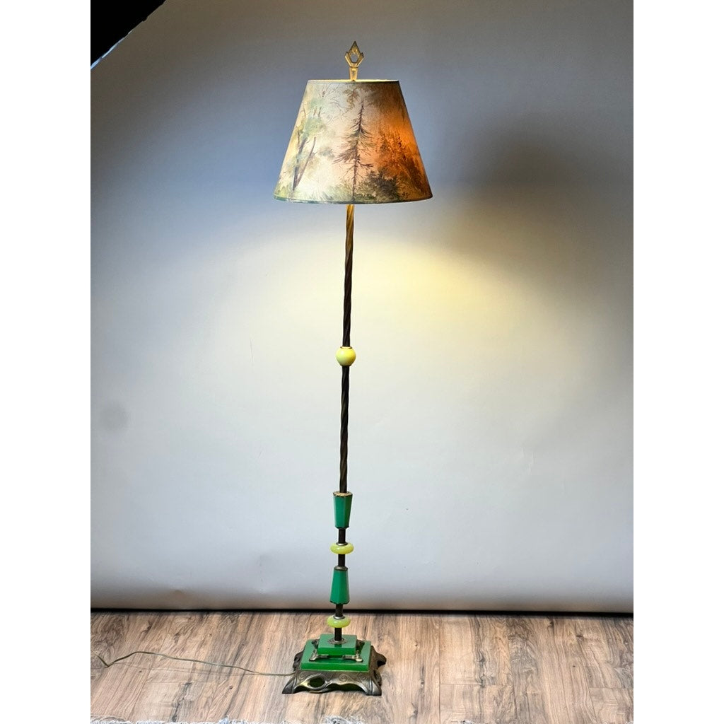 An Antique Art Deco Uranium and Slag Glass Lamp c. 1920 with a painted scenic shade and green and gold ornate base stands illuminated on a wood floor against a plain background, casting warm light. Brand: Unknown.