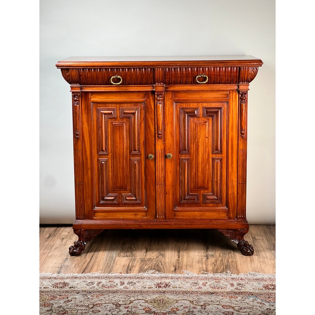 An Antique English Jacobean Revival Mahogany Buffet (c. 1915–1925) with ornate carving, two top drawers with brass handles, double doors below, and claw feet stands on a hardwood floor beside a patterned rug.