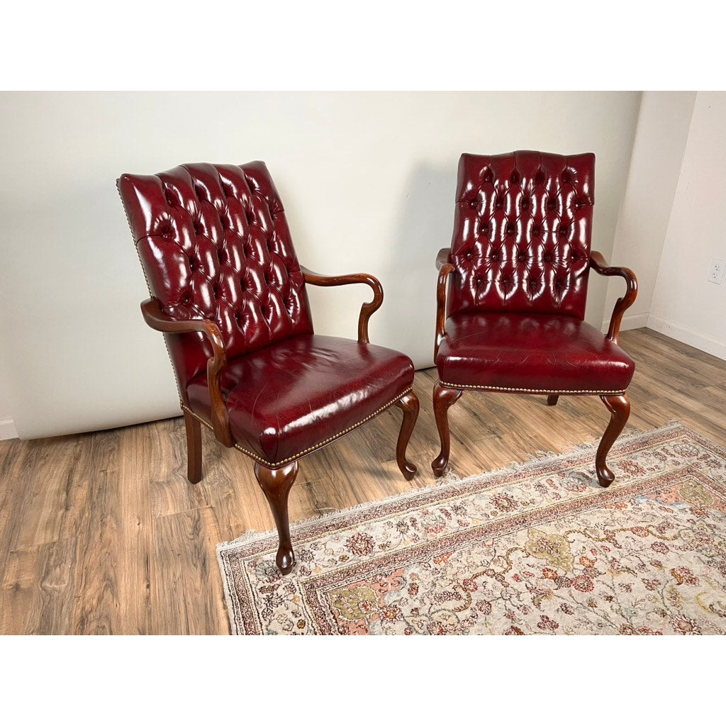 A Pair of Chippendale Style Tufted Burgundy Leather Library Armchairs, featuring classic wooden frames, are arranged on a wooden floor next to a patterned area rug and set against a light-colored wall.