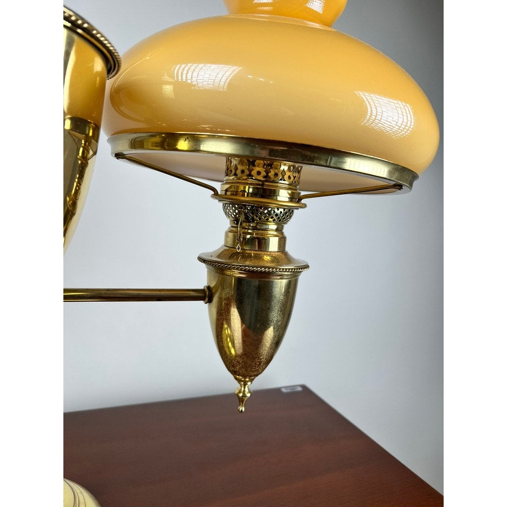 A close-up of a Bradley and Hubbard Antique Brass Double-arm Student Lamp with caramel glass shades, displayed on a wooden surface against a plain light gray background.