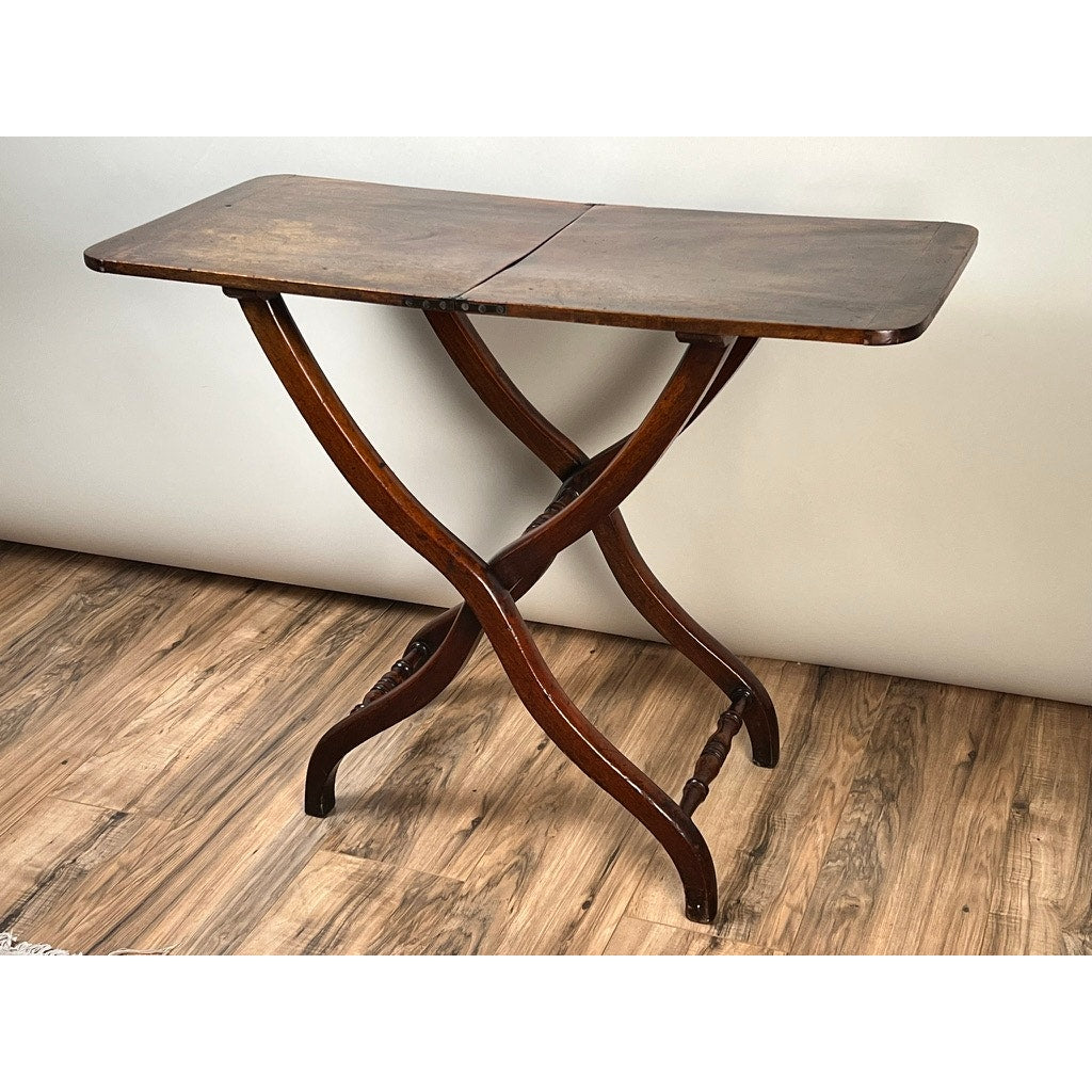 An Antique English Regency Mahogany Folding Coaching or Sporting Table (c. 1825–1845) with a rectangular top and crossed, curved legs sits on a wooden floor against a plain light wall, highlighting the elegance of this portable antique.
