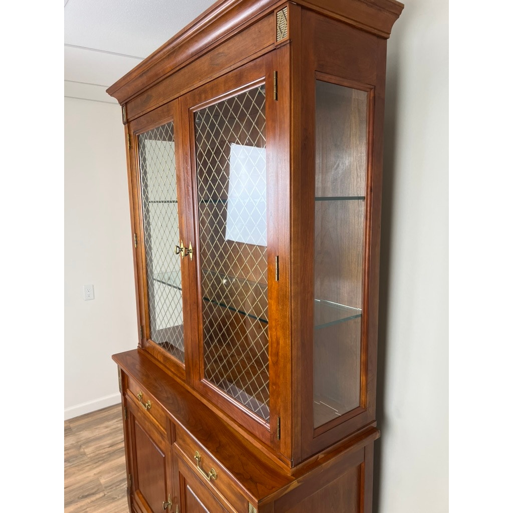 A Vintage William IV Style Cherry Glass Door Bookcase by Stickley stands against a white wall on a wood floor, featuring glass panel doors, wire mesh detailing, upper shelves, and lower drawers with brass handles.