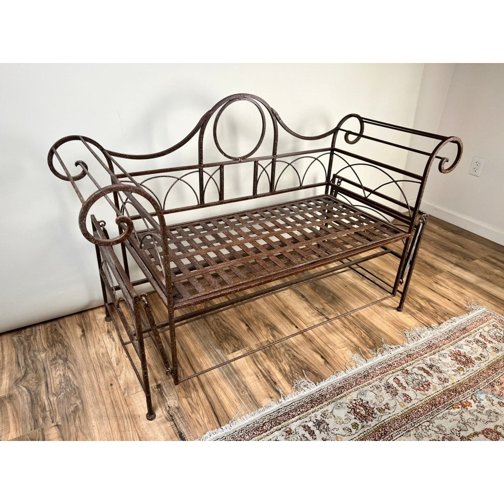 A Vintage Regency Style Wrought Iron Porch Glider with ornate scrollwork and a lattice seat sits on a wooden floor beside a patterned rug, set against a plain white wall. Brand: None/Unknown.