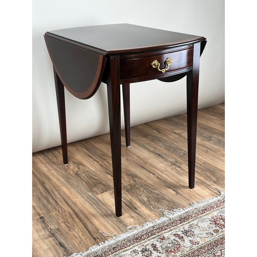 A Vintage Federal Style Edge Banded Mahogany End Table by Stickley with tapered legs and a single drawer with an ornate brass handle stands on a wooden floor near a patterned rug.
