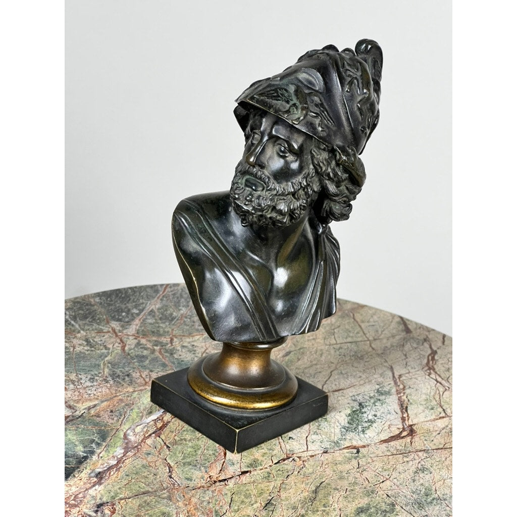 The Antique Small Bronze Bust of King Menelaus, c. Late 19th Century, features a bearded figure in an ornate helmet, turned slightly to the side and mounted on a square pedestal atop a marble-patterned table. Brand unknown.