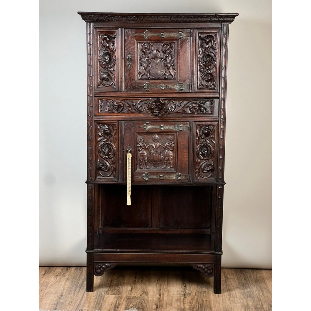 An Antique 19th-Century French Breton-style carved oak cabinet featuring deeply carved knights stands on a wooden floor against a plain wall, with two enclosed compartments and an open lower shelf.