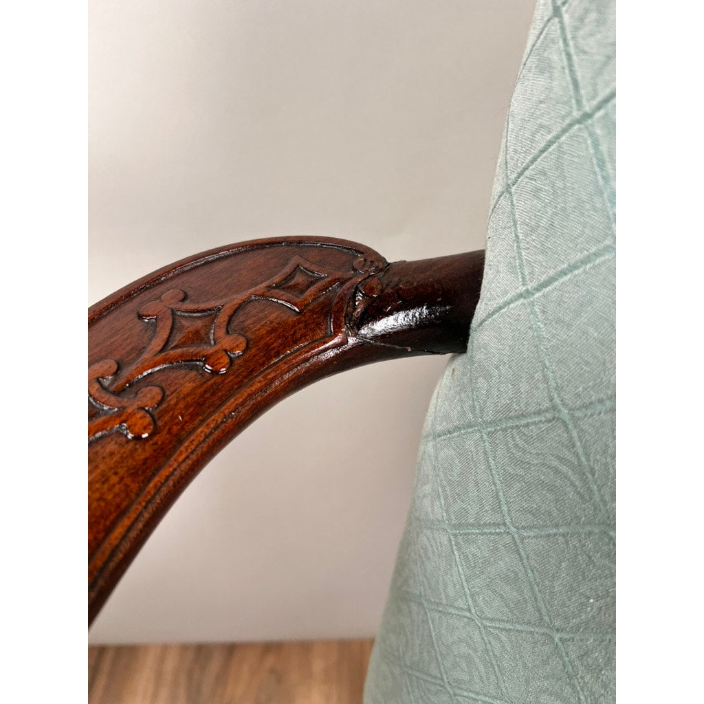 Close-up of an Antique George II Mahogany Gainsborough Open Arm Chair (mid-19th c.): wooden arm with intricate carving, attached to a padded, light green diamond-patterned backrest. Mahogany frame rests on wood floor against a neutral background.
