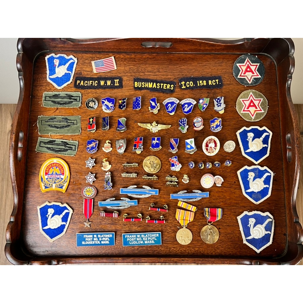 A wooden tray displays an attributed WWII 158th Infantry Bushmasters grouping for Frank W. Slatcher, Company I, Pacific Theater, featuring military badges, medals, patches, star pins, ribbon medals, and nameplates.