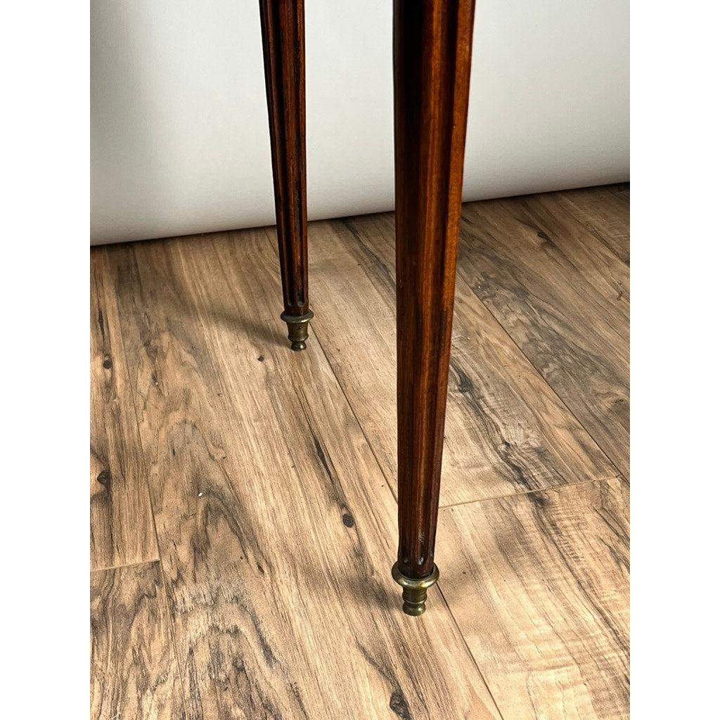 Close-up of two slender mahogany legs with brass caps from an antique 19th-century French Louis XVI-style Bouillotte table with a marble top and pierced brass gallery, shown on a softly lit light brown wood floor. Brand: Unknown.