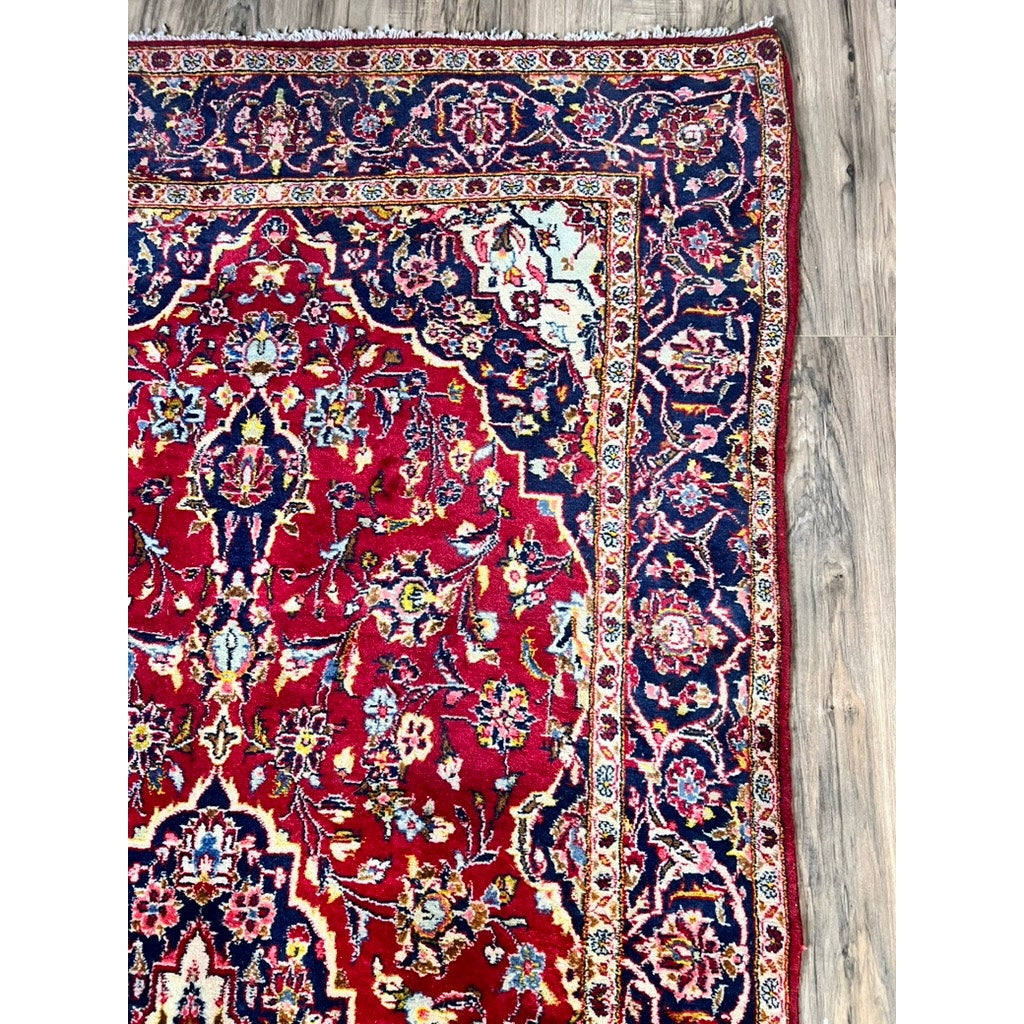 A close-up of a Vintage Hand-Knotted Persian Kashan Wool Gallery Rug (c. 1950–1970, 4’5 x 8’8) shows its intricate floral patterns in red, blue, yellow, and white on a wooden floor, with detailed borders and vibrant colors. Brand: Unknown.