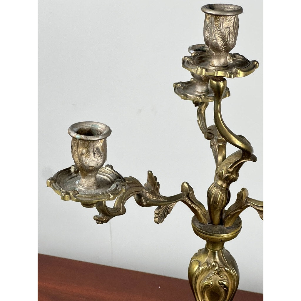A close-up of a Vintage Brass Louis XV Style Four Light Candelabra with ornate details, displayed on a wooden surface against a plain white wall. Brand: None and/or Unknown.