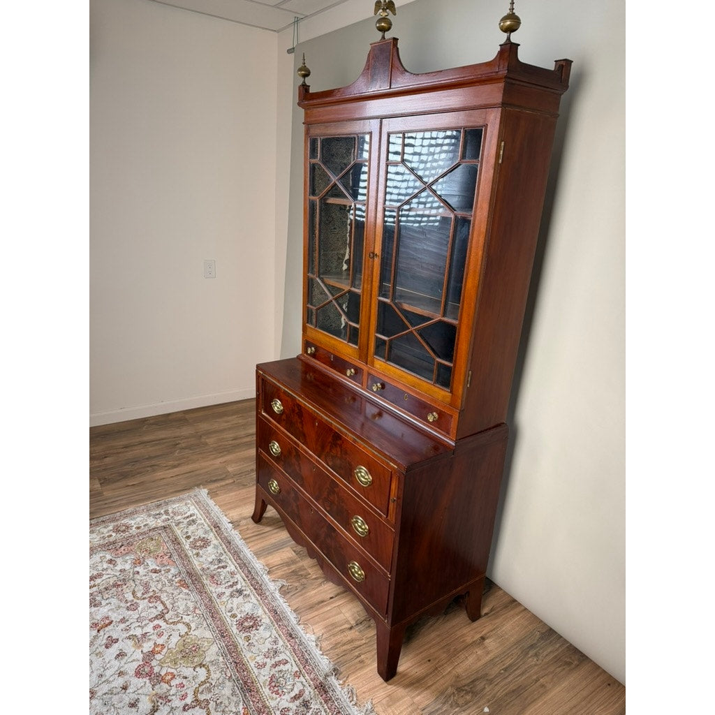 A Federal Flame Mahogany Secretary Bookcase with Eagle Finial, dating from circa 1810–1825, stands against a wall on a wooden floor beside a patterned rug. Featuring upper glass doors, brass handles, and three drawers below, this elegant piece reflects the refined design of early 19th-century American cabinetmaking during the Federal period.