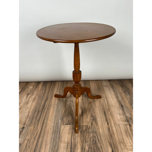The Antique Tiger Maple Queen Anne Style Candle Stand by Pearl City Antiques, dating to the mid 19th century, features a small, round wooden pedestal design with a turned central column and three elegantly curved legs. Crafted from tiger maple, this piece sits gracefully on a wood-patterned floor against a plain light-colored wall, echoing the charm of antique candle stands.