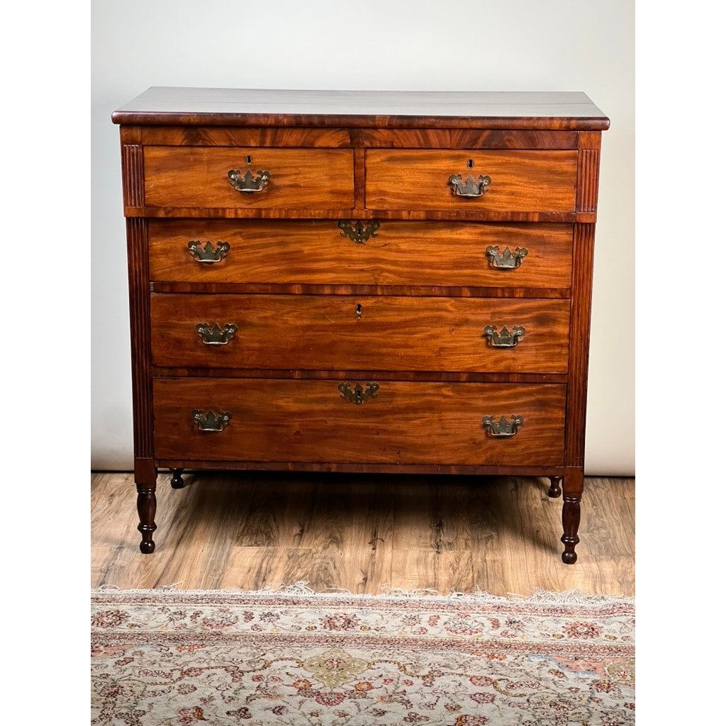 An Antique George III Sheraton Flame Mahogany Chest of Drawers from the early 19th century, featuring five drawers with ornate metal handles and turned legs, stands polished in rich brown on a hardwood floor beside a patterned rug.