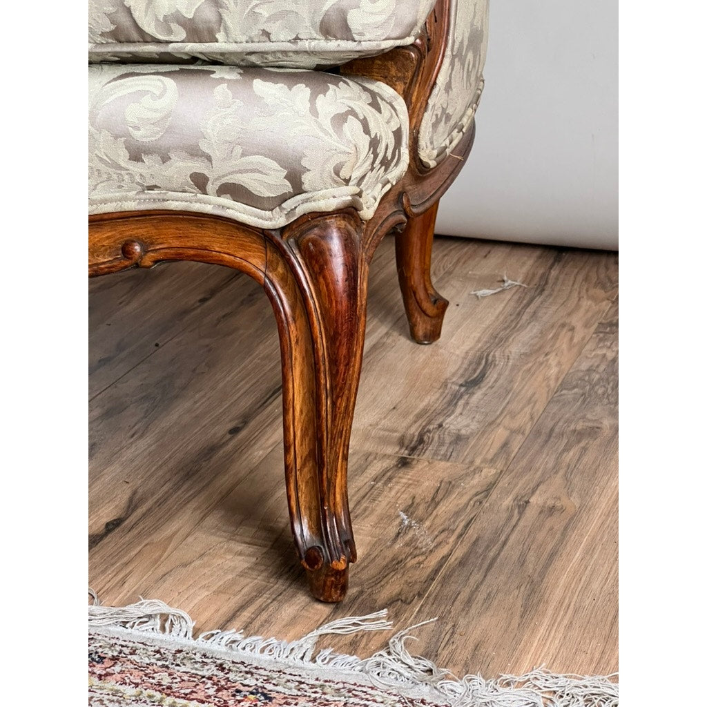 Close-up of an Antique French Louis XV Rococo Style Bergère Arm Chair, c. Mid 19th Century, featuring carved walnut legs and newer damask upholstery in beige and cream floral pattern, on a wooden floor beside a fringed rug. Brand: Unknown.