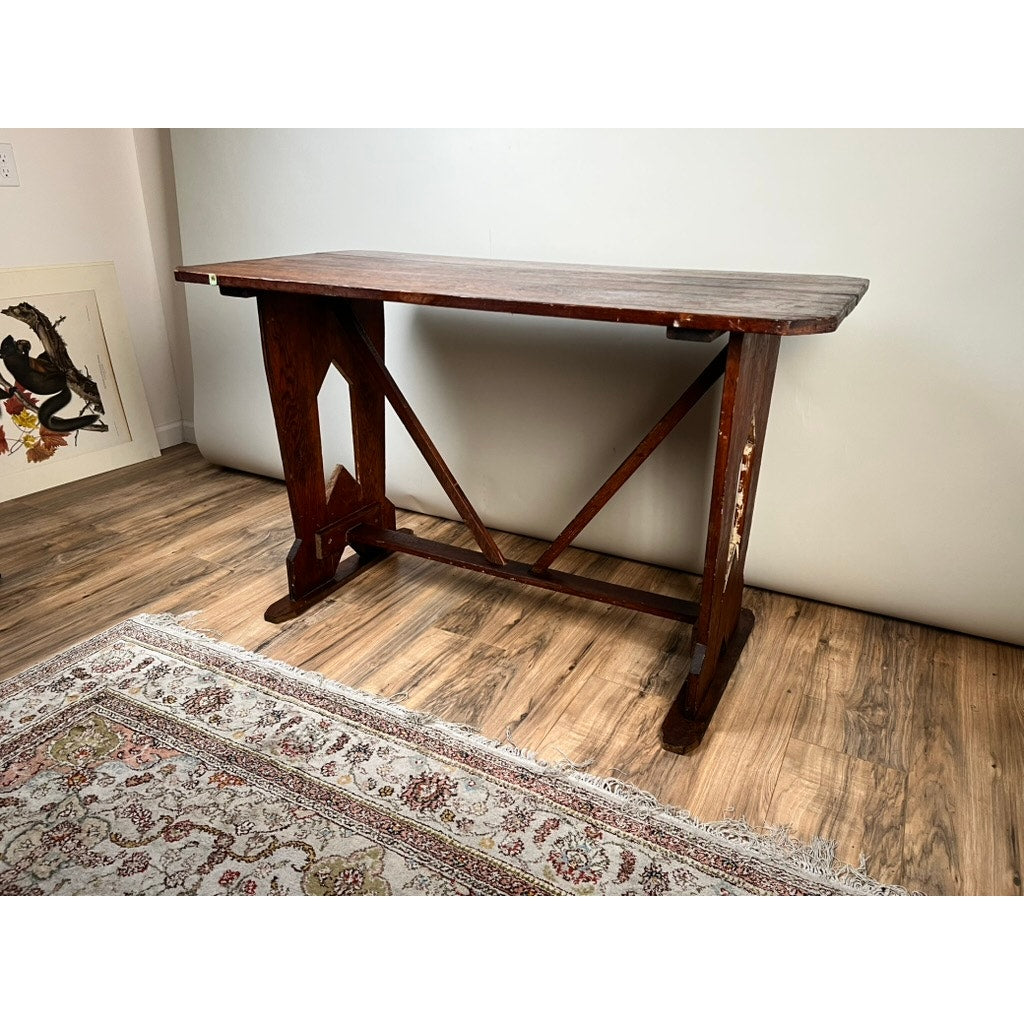 The Antique Rustic Italian Fratino Trestle Table (late 19th century) stands on a wooden floor by a patterned rug and framed artwork leaning on a white wall, ideal for farmhouse décor enthusiasts.