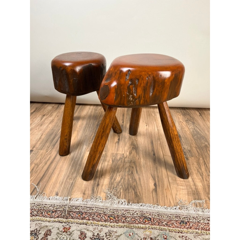 A pair of vintage live edge Wabi Sabi cherry stools, signed by Neil Panyik, with round seats and three angled legs each, sit on a wooden floor next to a patterned rug, highlighting their natural and artisanal charm.