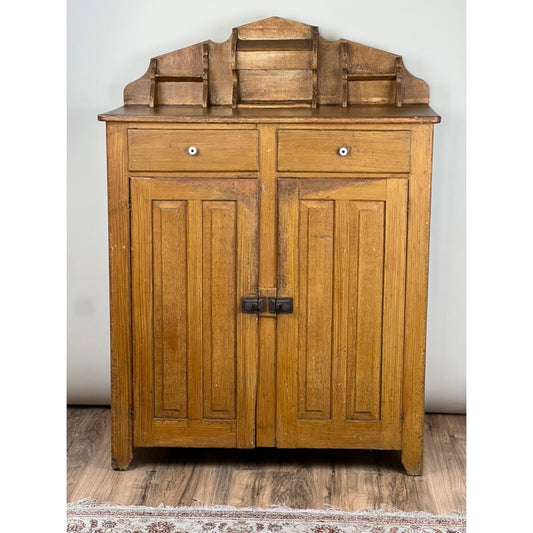 The Antique Victorian Country Grain Painted Jelly Cupboard c. 1870 by Pearl City Antiques features two paneled doors, two small upper drawers with black metal handles, a decorative top shelf, and stands on a wood floor with part of a patterned rug visible.