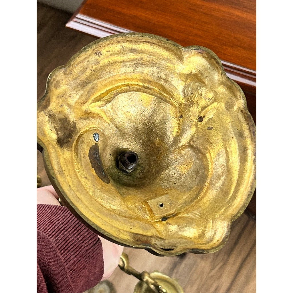 Close-up of the underside of a Vintage Brass Louis XV Style Four Light Candelabra with ornate details and hollow center, held by a person in a maroon sleeve. A wooden surface with white trim is visible in the background. Brand: Unknown.